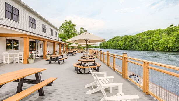 A deck with picnic tables and chairs overlooking a river, with a building on one side. - The Cove at Sylvan Beach - Pet friendly lakeside cottage with a pontoon boat! (Sylvan Beach)