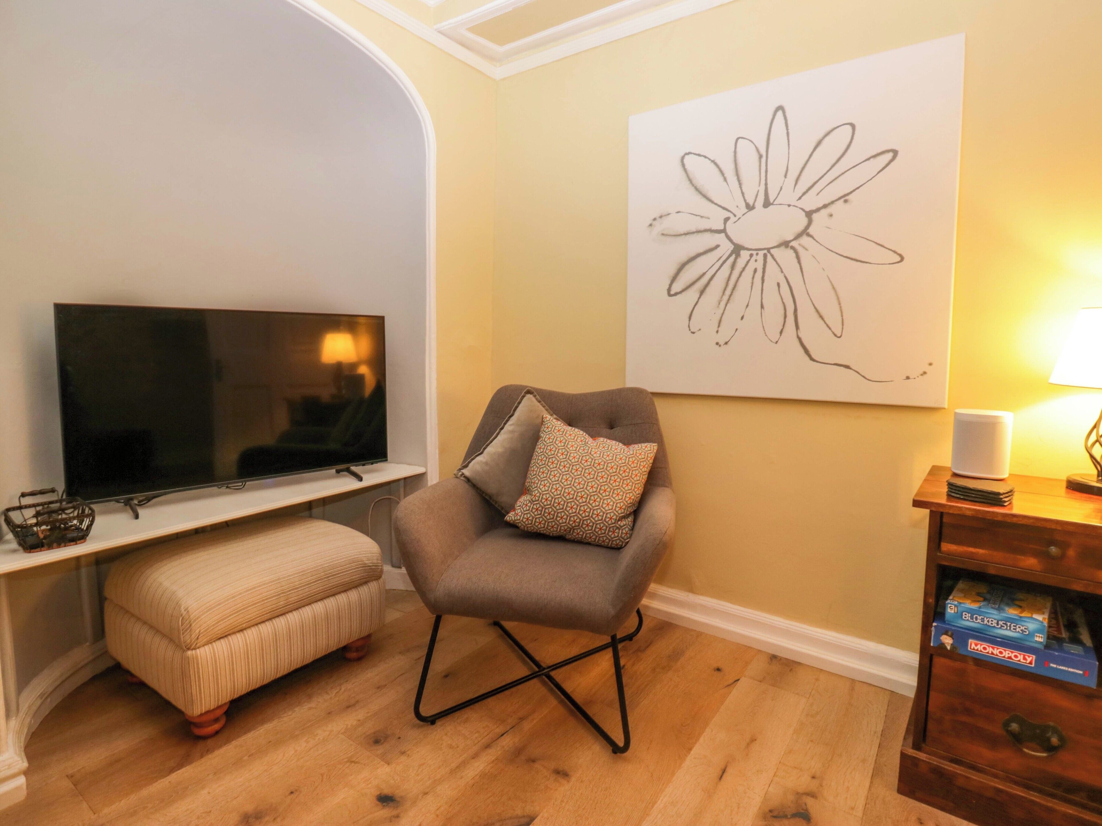 A living room with a flat-screen TV, a grey armchair with patterned cushions, a small ottoman, and a wooden side table with a lamp and board games.