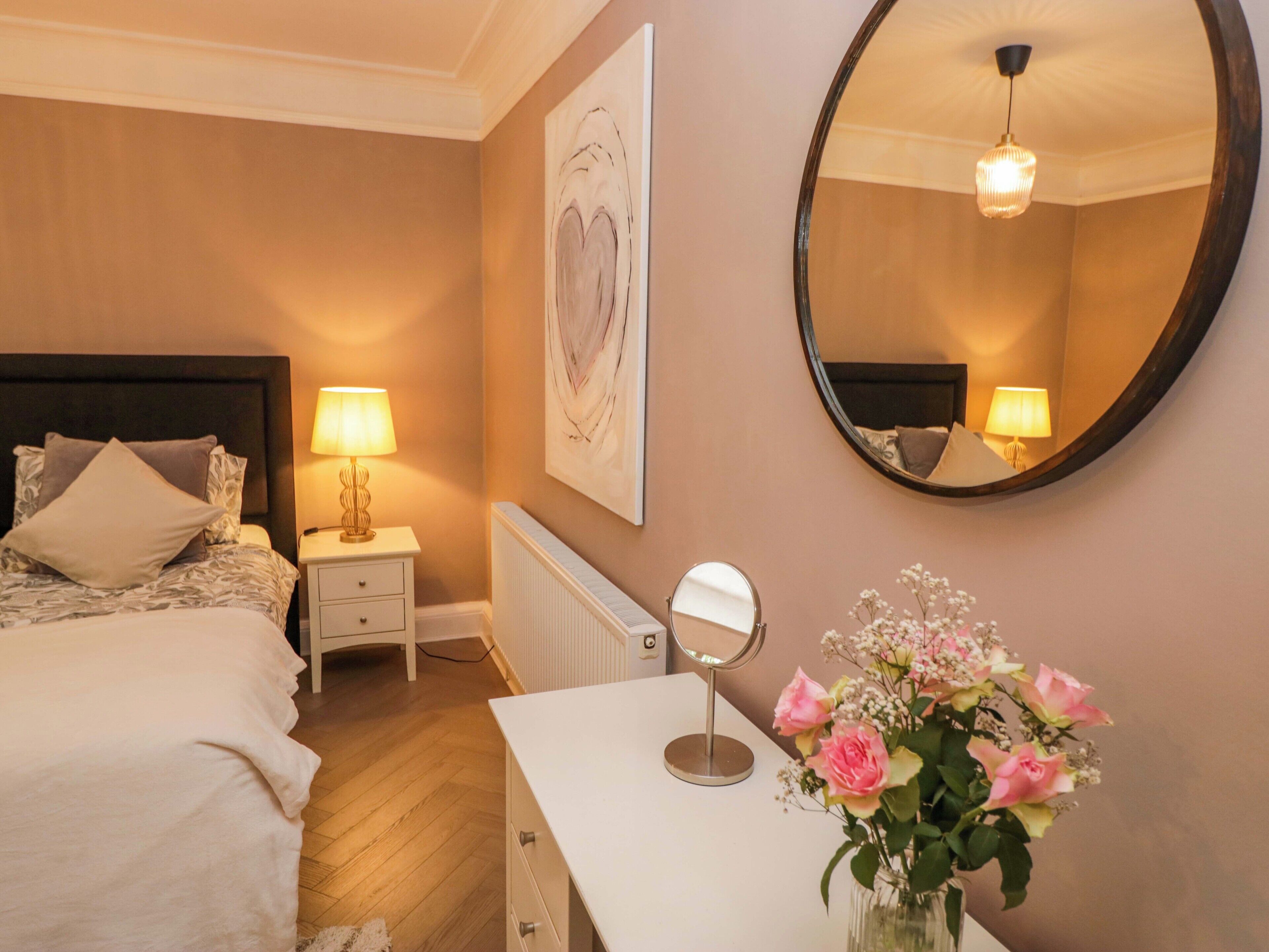 A bedroom with a bed, bedside table, lamp, mirror, and a vase with flowers.