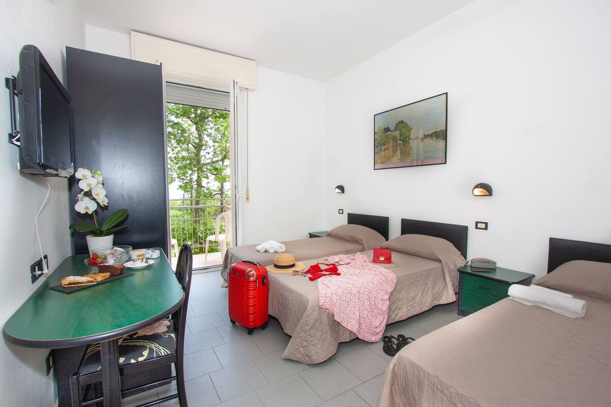 Ein Hotelzimmer mit zwei Betten, einem Schreibtisch mit Stuhl, einem Fernseher, einem Esstisch mit Pflanze und einem Balkon mit Blick auf Bäume.