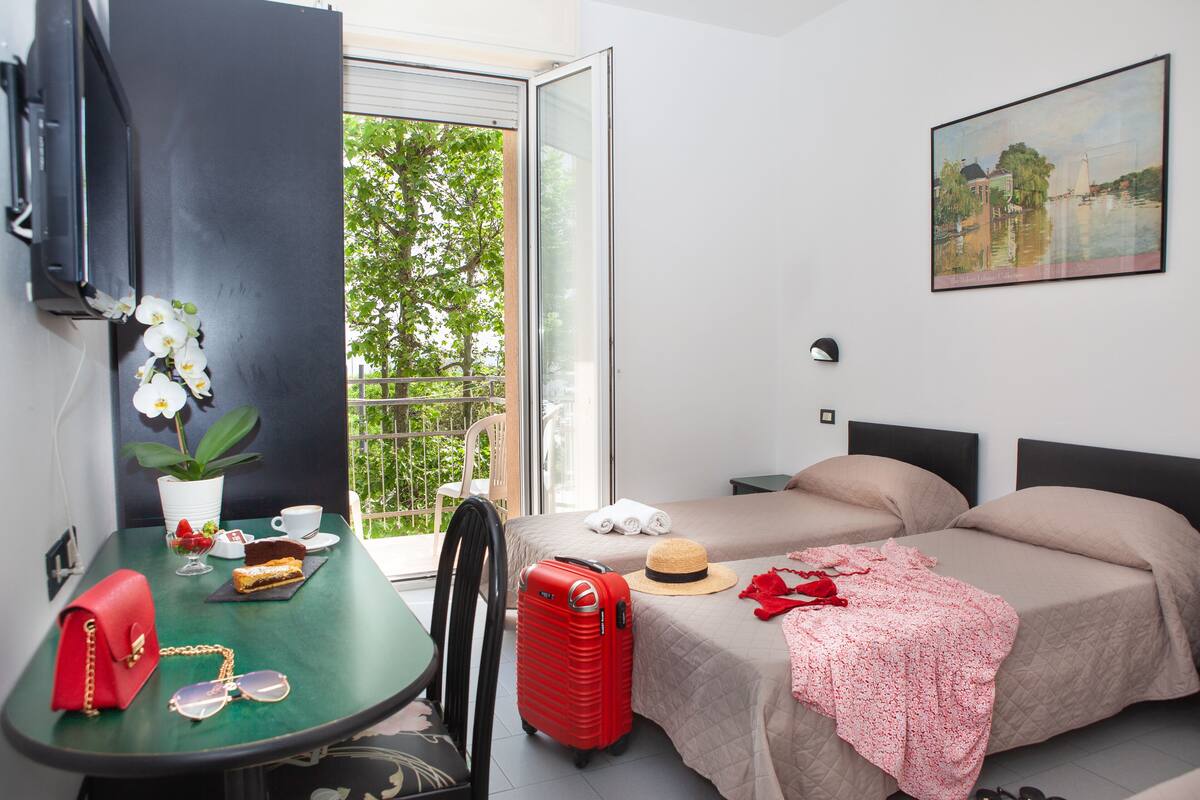 Ein Hotelzimmer mit Bett, Tisch, Stuhl, Koffer, Hut und einem Bild an der Wand.