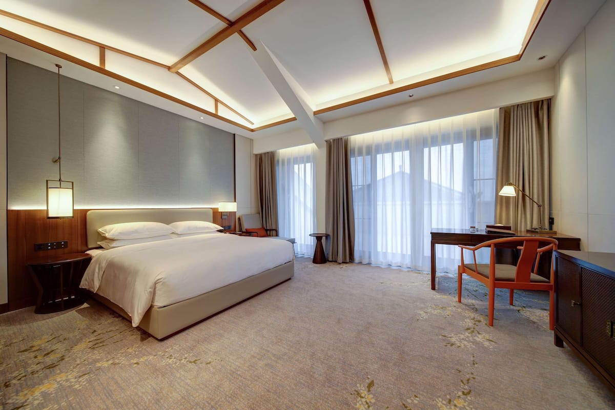 Ein Hotelzimmer mit einem großen Bett, einem Schreibtisch mit Stuhl, einer Lampe und Blick ins Freie durch das Fenster.