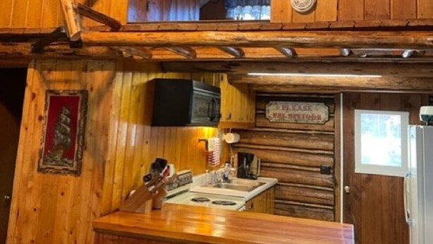 A rustic cabin interior with wooden walls, a lofted sleeping area, a kitchen with basic amenities, and a wooden chair. - Wrangler Log Cabin Sleep 8 (Condon)