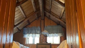 A cabin with a sloped wooden ceiling, two windows with curtains, a wooden dresser, and a bed. - Wrangler Log Cabin Sleep 8 (Condon)