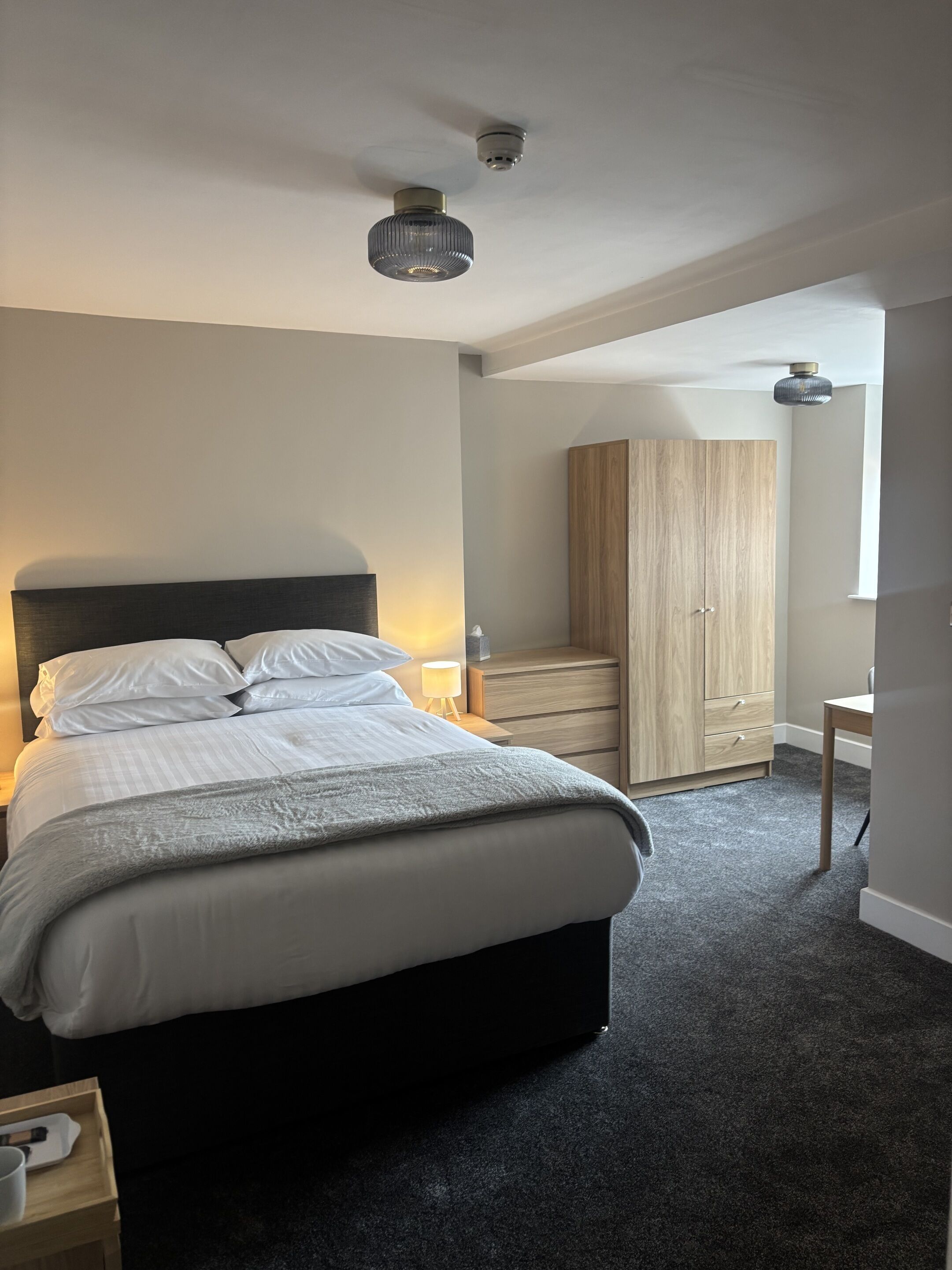 A bedroom with a bed, bedside table, lamp, wardrobe, and a desk with a chair.