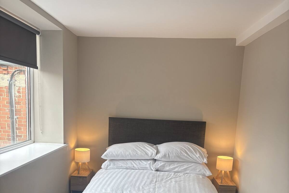 Een net opgemaakt bed met witte lakens, twee bedlampjes en een raam met uitzicht op een bakstenen muur.