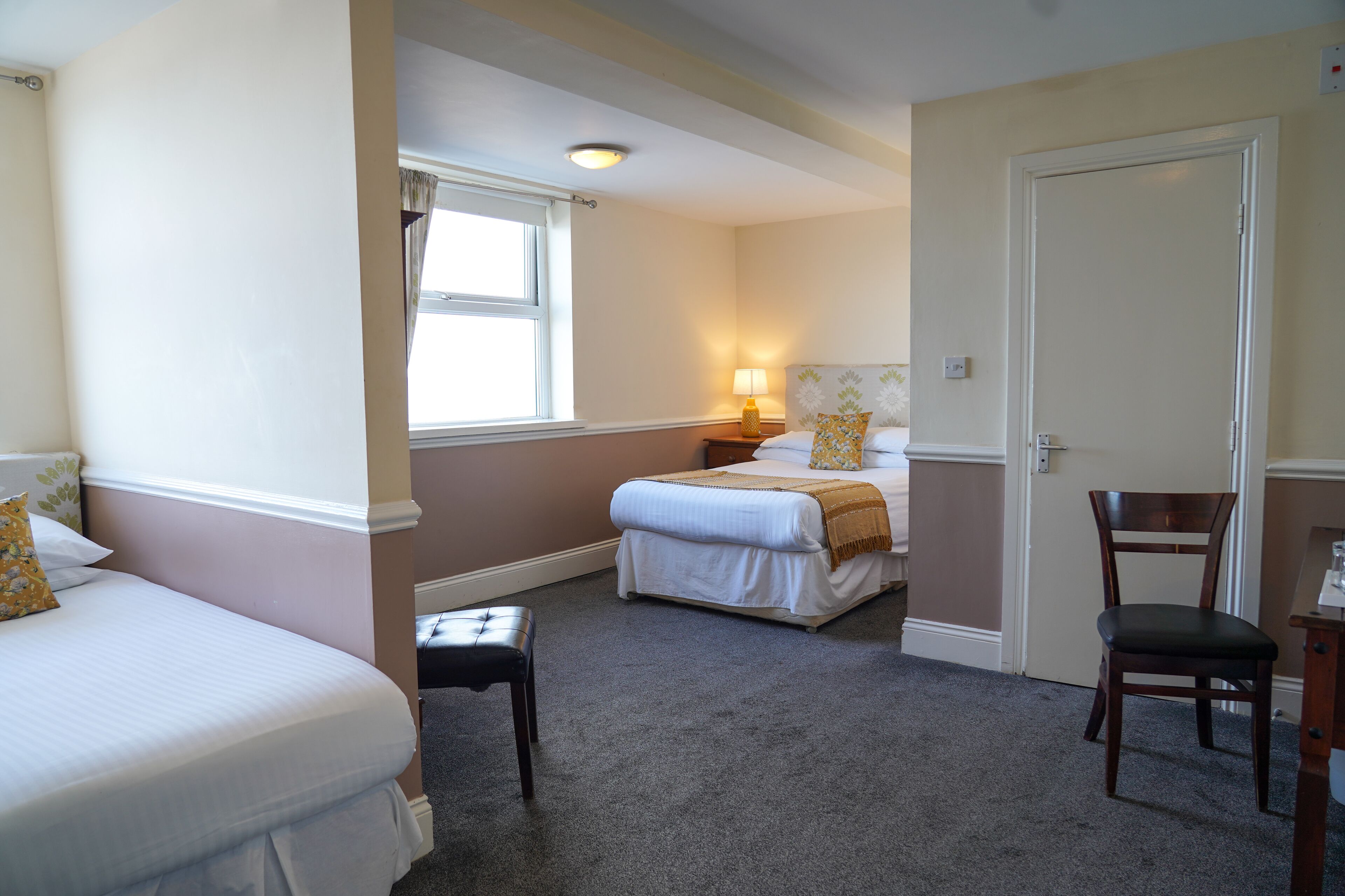 A hotel room with two beds, a chair, a nightstand with a lamp, and a window.