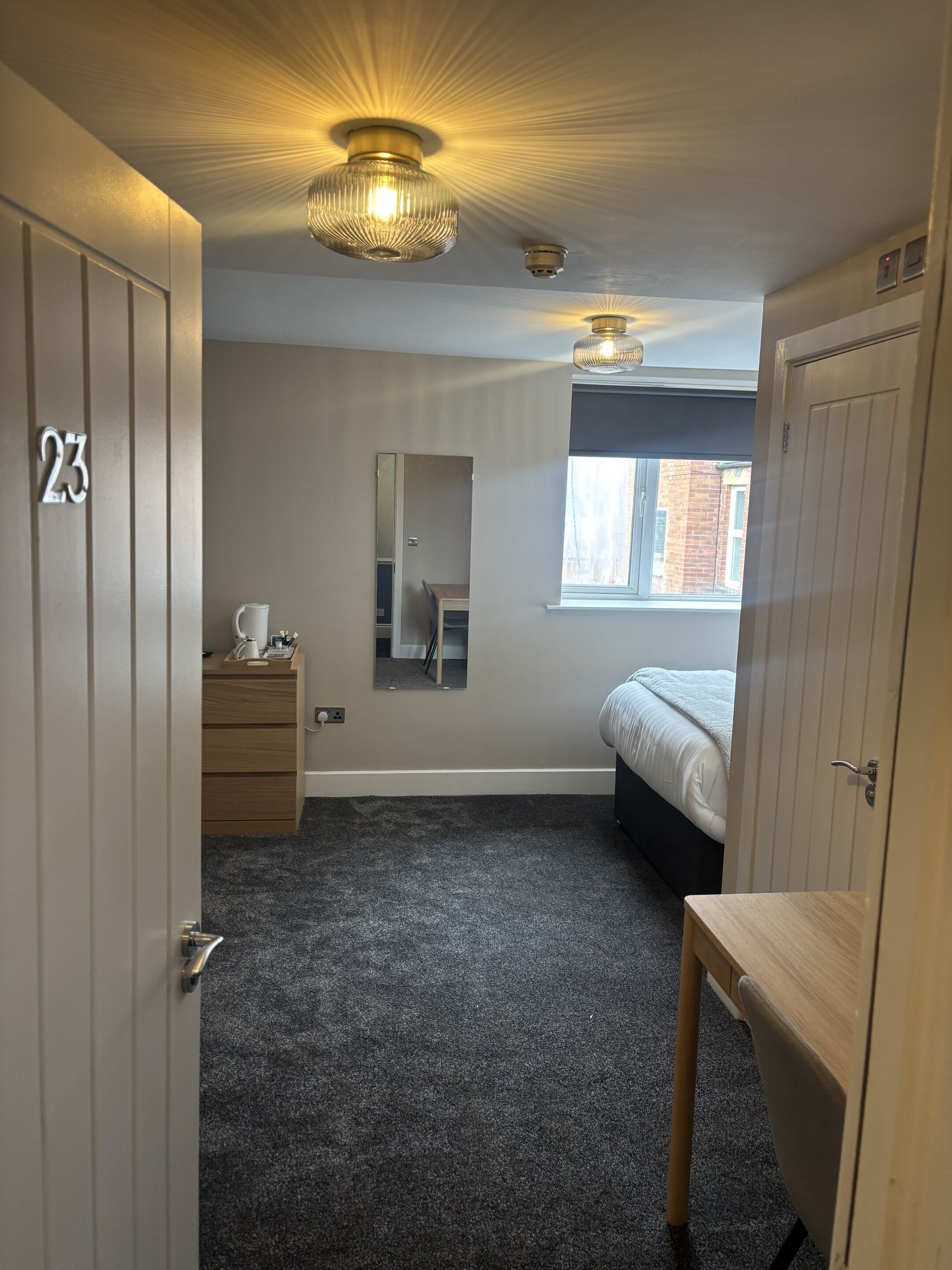 Een slaapkamer met een bed, een kaptafel, een spiegel, een raam en een deur met de tekst '23'.