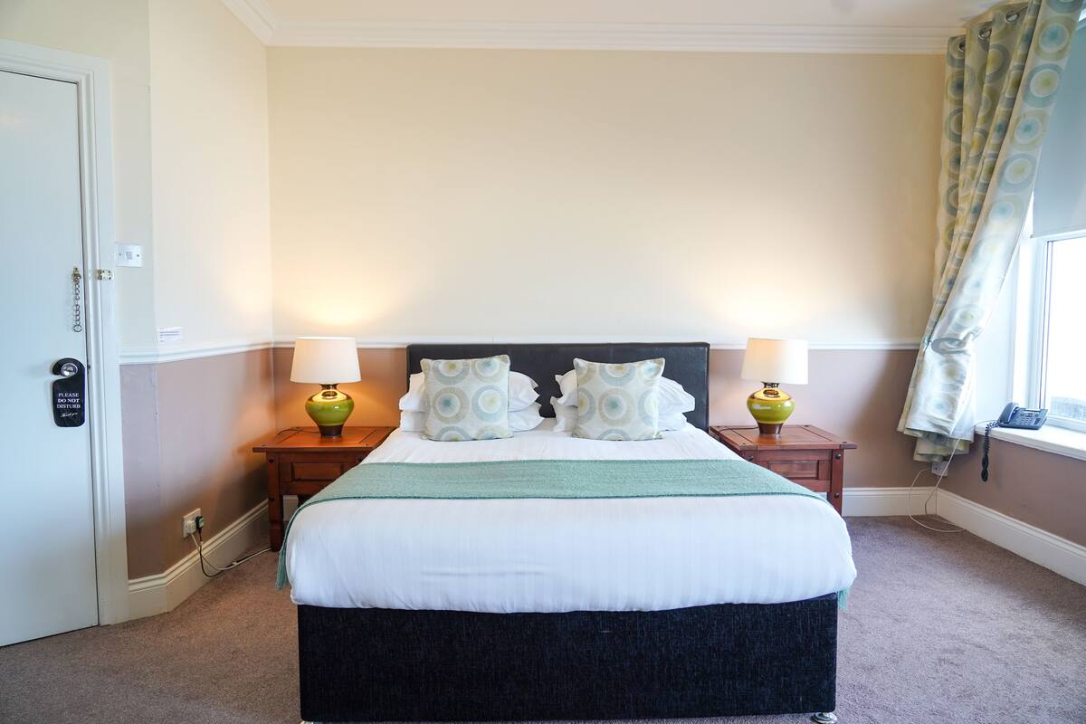 Een net opgemaakt bed met een wit- en groenbedspreider, twee bijpassende kussens en twee nachtlampjes.