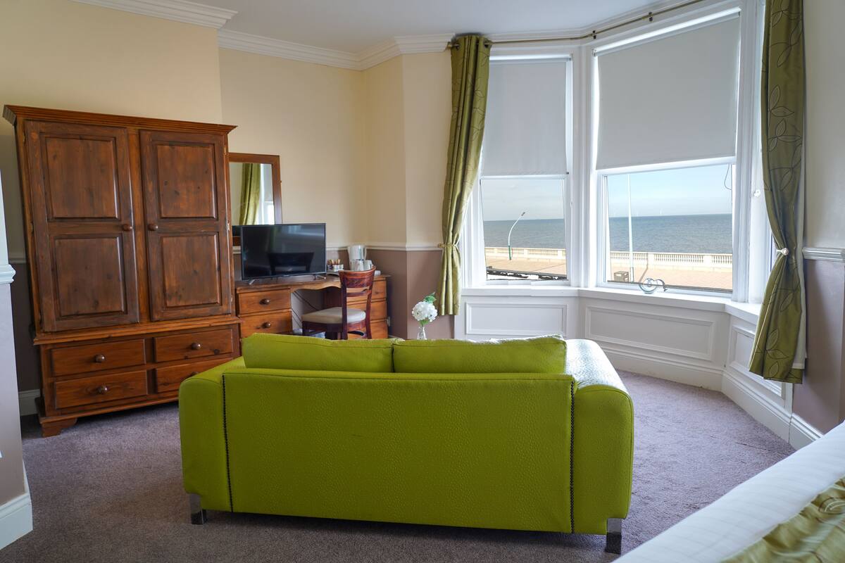 Een kamer met een groene bank, een houten garderobe, een televisie en uitzicht op de zee door een groot raam.
