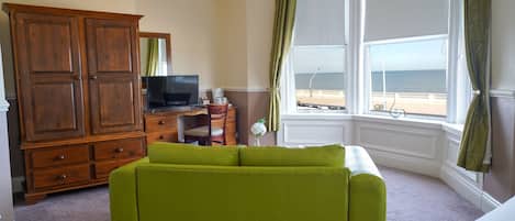 A room with a green sofa, wooden wardrobe, television, and a view of the sea through a large window.