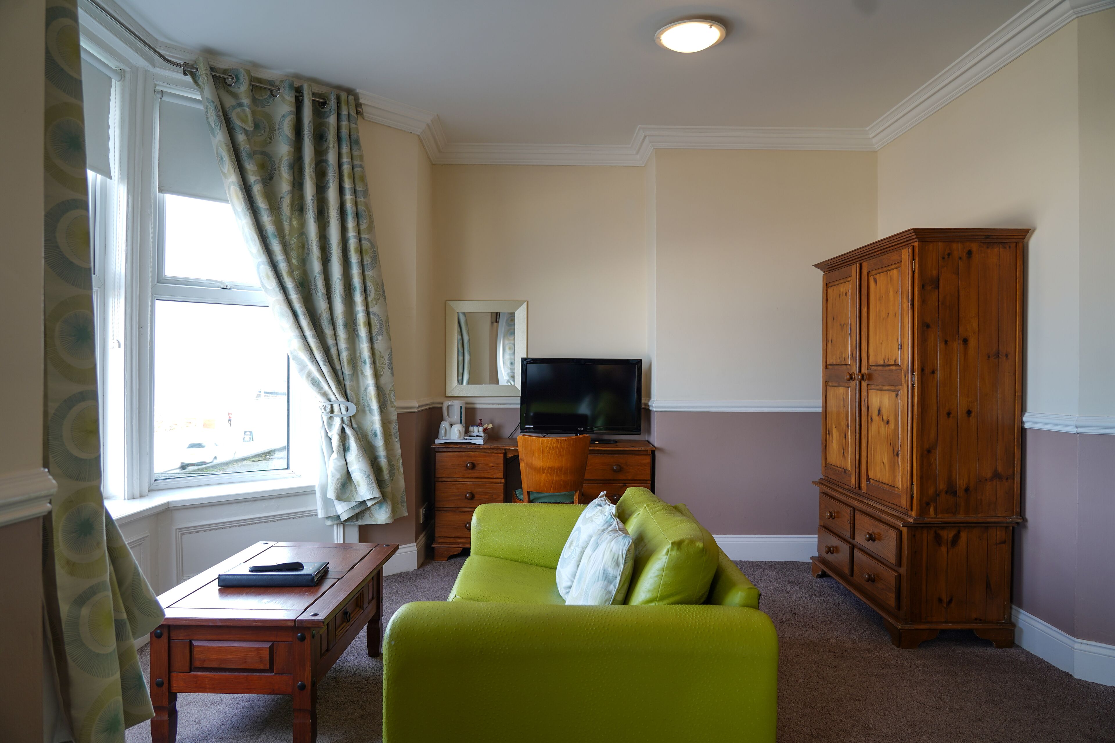 A living room with a green sofa, a wooden cabinet, a television on a wooden stand, and a window with curtains.