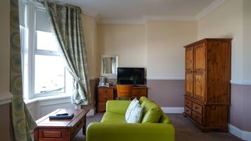 A living room with a green sofa, a wooden cabinet, a television on a wooden stand, and a window with curtains.