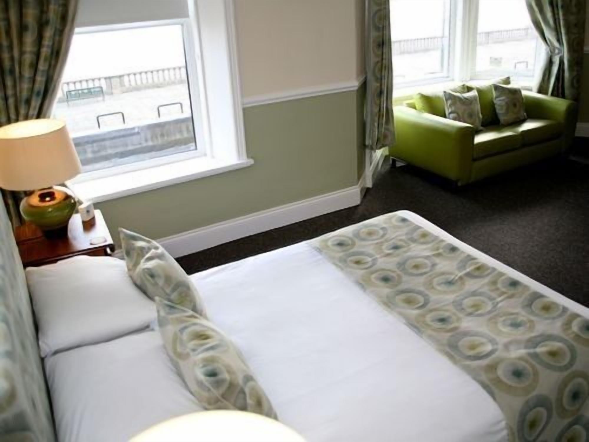 Una habitación de hotel con una cama, una mesita de noche con lámpara, una ventana con cortinas y un sofá verde en la esquina.