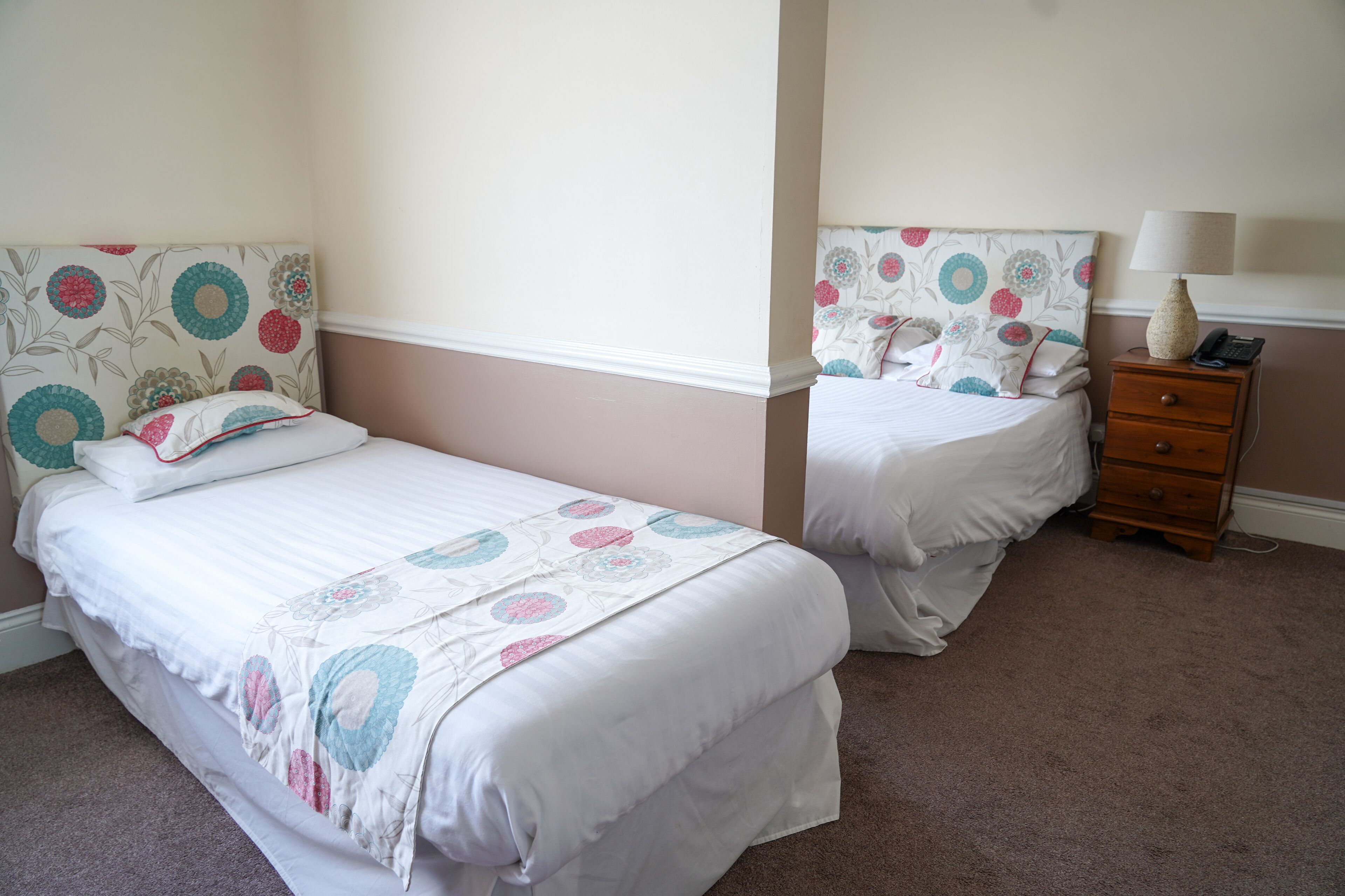 A bedroom with two beds, each with floral headboards and white bedding. There is a wooden nightstand with a lamp and a telephone.