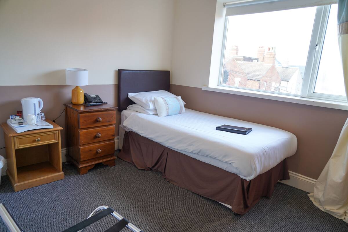 Een hotelkamer met een eenpersoonsbed, een nachtkastje met een lamp, een telefoon en een raam met uitzicht op gebouwen.