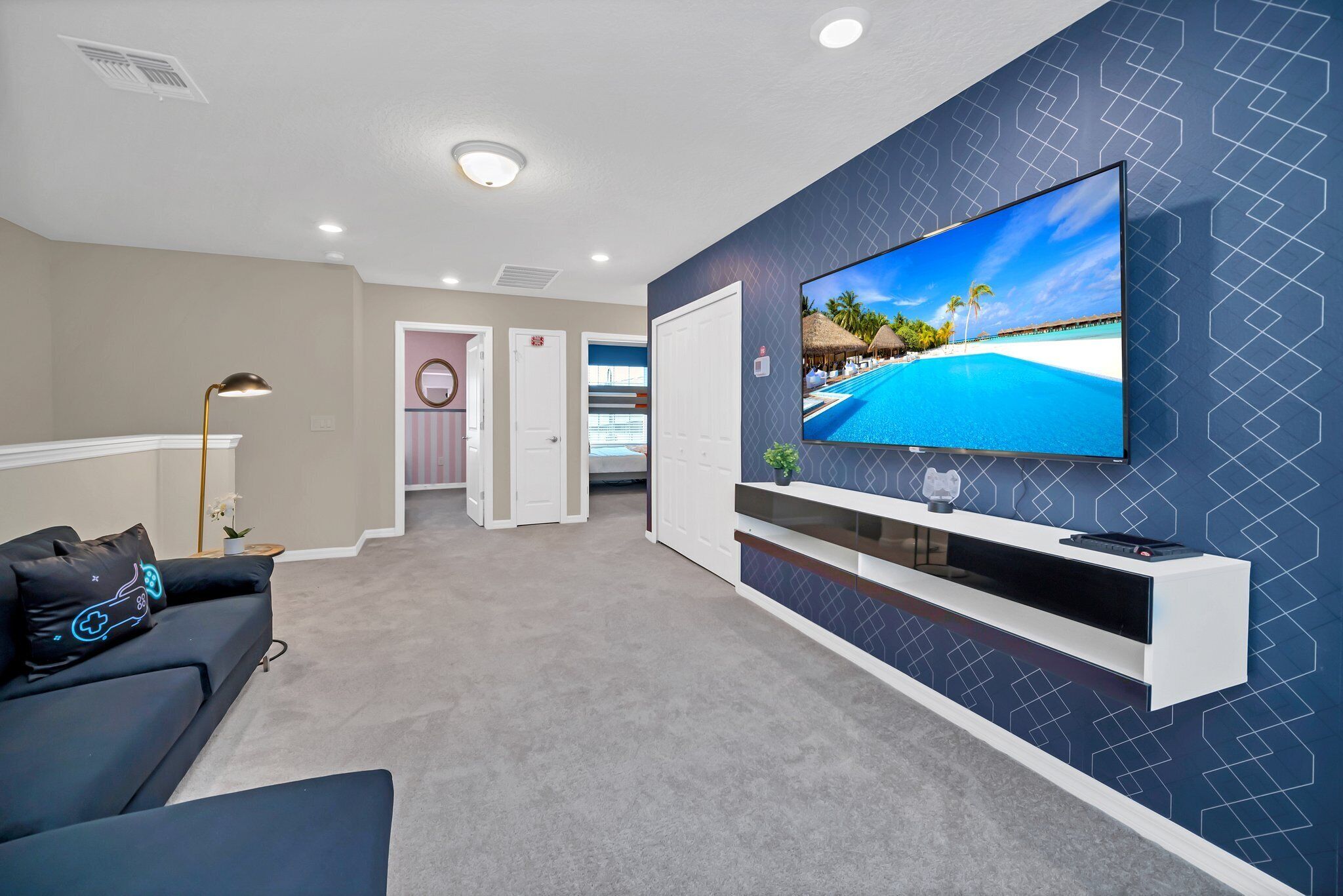 A modern living room with a large flat-screen TV mounted on a wall, a dark-colored sofa with decorative pillows, a side table with a lamp, and a hallway leading to a door.