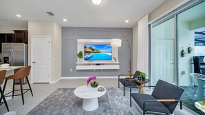 A modern living room with a flat-screen TV, a dining area with chairs, a coffee machine, and a kitchen area with a refrigerator. - Free Waterpark Private Splash Pool Near Disney (Davenport)
