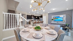 A modern dining area with a round table set for six, a staircase with white railings, and a kitchen in the background. - Free Waterpark Private Splash Pool Near Disney (Davenport)