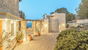 A stone building with a paved courtyard, potted plants, and a view of the sea. - Thomais Villas Estate | Sea View & Private Pool (Agia Pelagia)