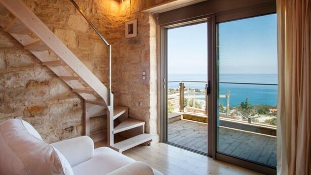 A room with a stone wall, a white staircase, a white sofa, and a balcony with a view of the sea. - Thomais Villas Estate | Sea View & Private Pool (Agia Pelagia)