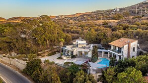 A modern villa with a swimming pool, stone architecture, and a view of the surrounding hills. - Thomais Villas Estate | Sea View & Private Pool (Agia Pelagia)