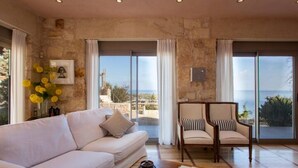 A living room with a white sofa, a wooden chair, and a view of the sea through large windows. - Thomais Villas Estate | Sea View & Private Pool (Agia Pelagia)