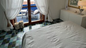 Ein Hotelzimmer mit einem Bett, einem an der Wand montierten Fernseher, einem Fenster mit weißen Vorhängen und einem gefliesten Boden.