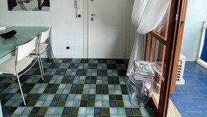 Ein kleiner, kompakter Raum mit Fliesenboden, einer weißen Tür, einer weißen Klimaanlage, einem Tisch mit Stühlen und einem Fenster mit Vorhängen.