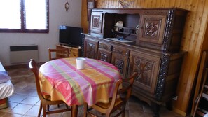 Une pièce chaleureuse avec un meuble en bois, une télévision, une table recouverte d’une nappe colorée et une chaise.