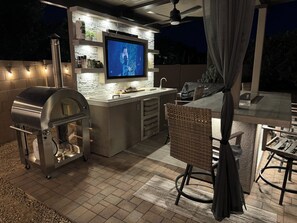 An outdoor kitchen with a grill, a TV, and a bar area. - Luxury 4 bed, 4 bath with Pool & Spa 🌞 Summer Special (Goodyear)