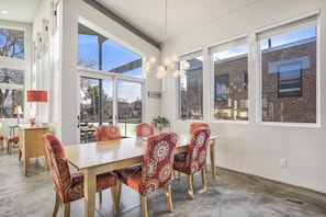 Un espace repas comprenant une grande table et six chaises, de grandes fenêtres et un luminaire suspendu.