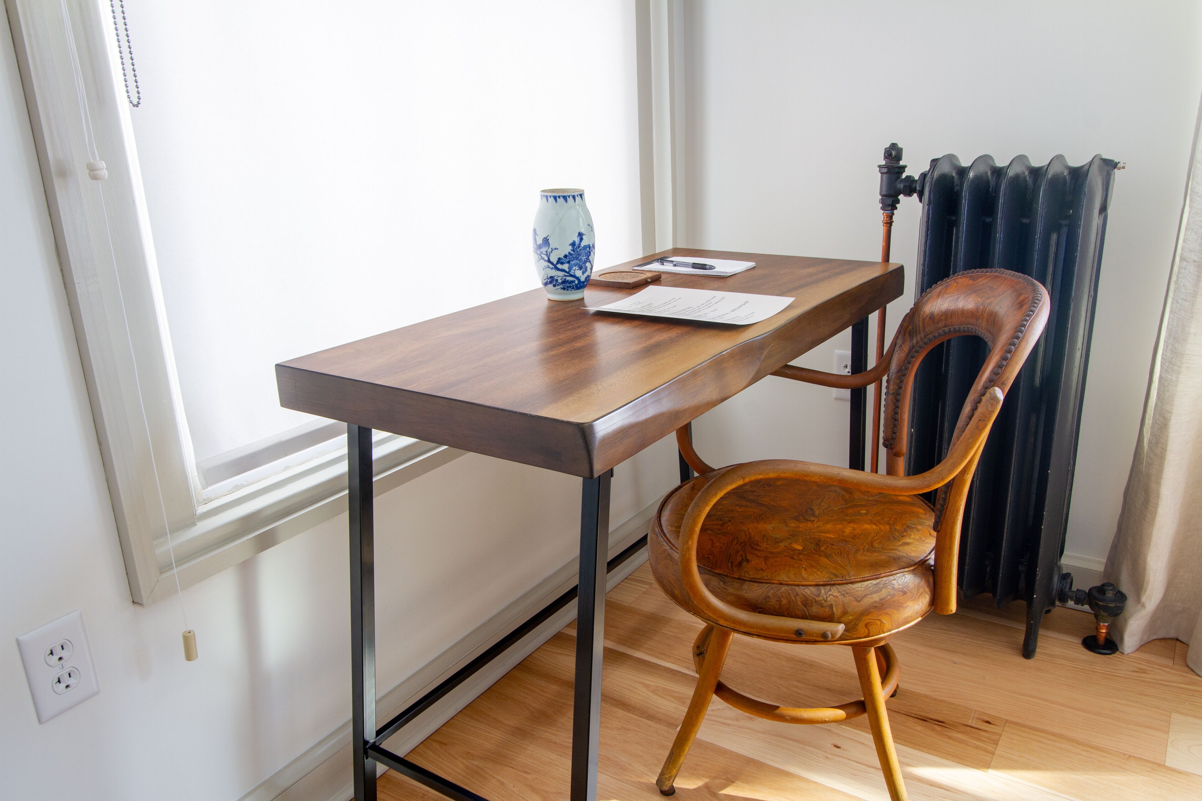 A wooden table with a vase and papers, a wooden chair, and a radiator in a room with a window and electrical outlet.