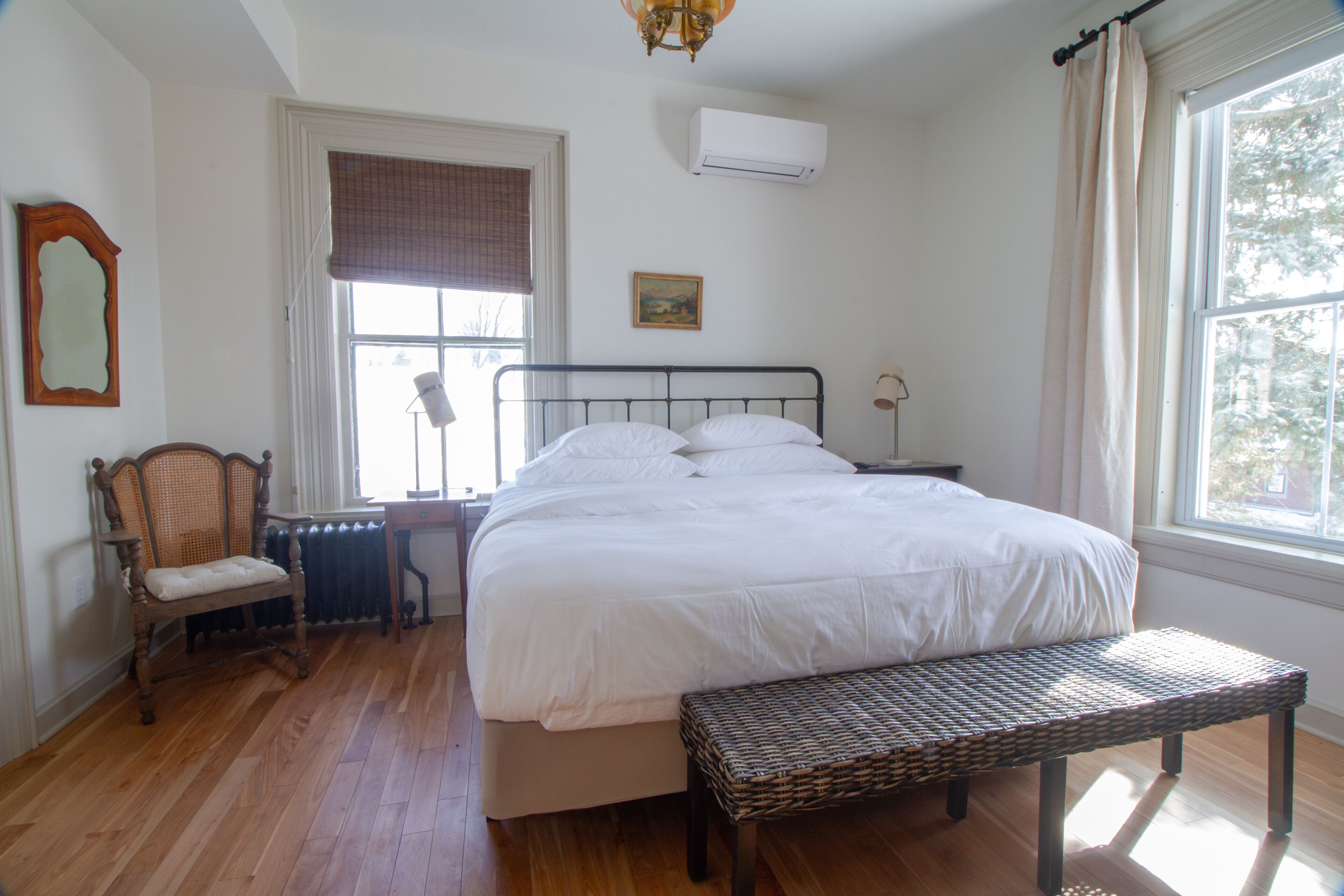 Un dormitorio con una cama grande, una silla de madera, un banco de mimbre, un radiador y una ventana con cortinas.