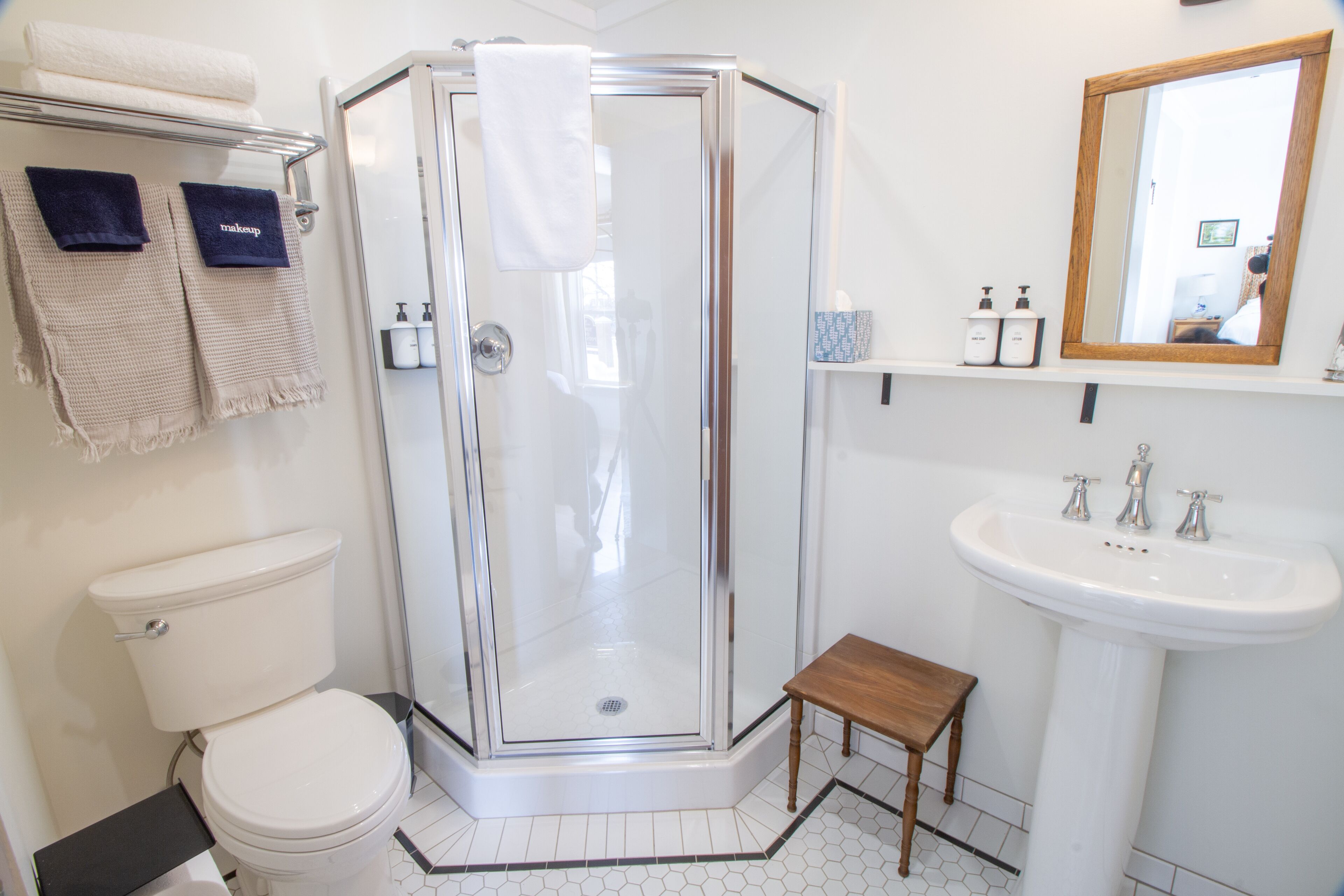 Een badkamer met een douchecabine van glas, toilet, wastafel en een klein houten krukje.