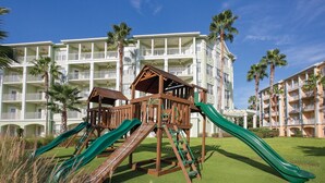 Un parque infantil con toboganes y estructuras de escalada frente a un hotel con múltiples balcones y palmeras.