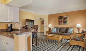 A hotel room with a kitchenette, a sofa set, a coffee table, and a striped carpet.