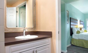Un baño con lavamanos, espejo y una puerta que da a un dormitorio con cama y mesita de noche.