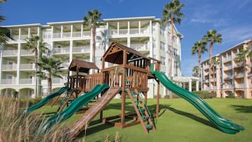 A playground with slides and climbing structures in front of a hotel with multiple balconies and palm trees.