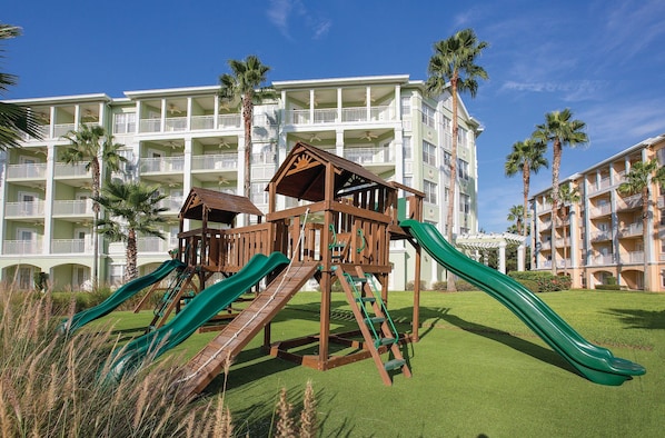 A playground with slides and climbing structures in front of a hotel with multiple balconies and palm trees.