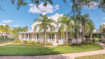 A large, white colonial-style house with a wrap-around porch, surrounded by well-maintained lawns and palm trees.