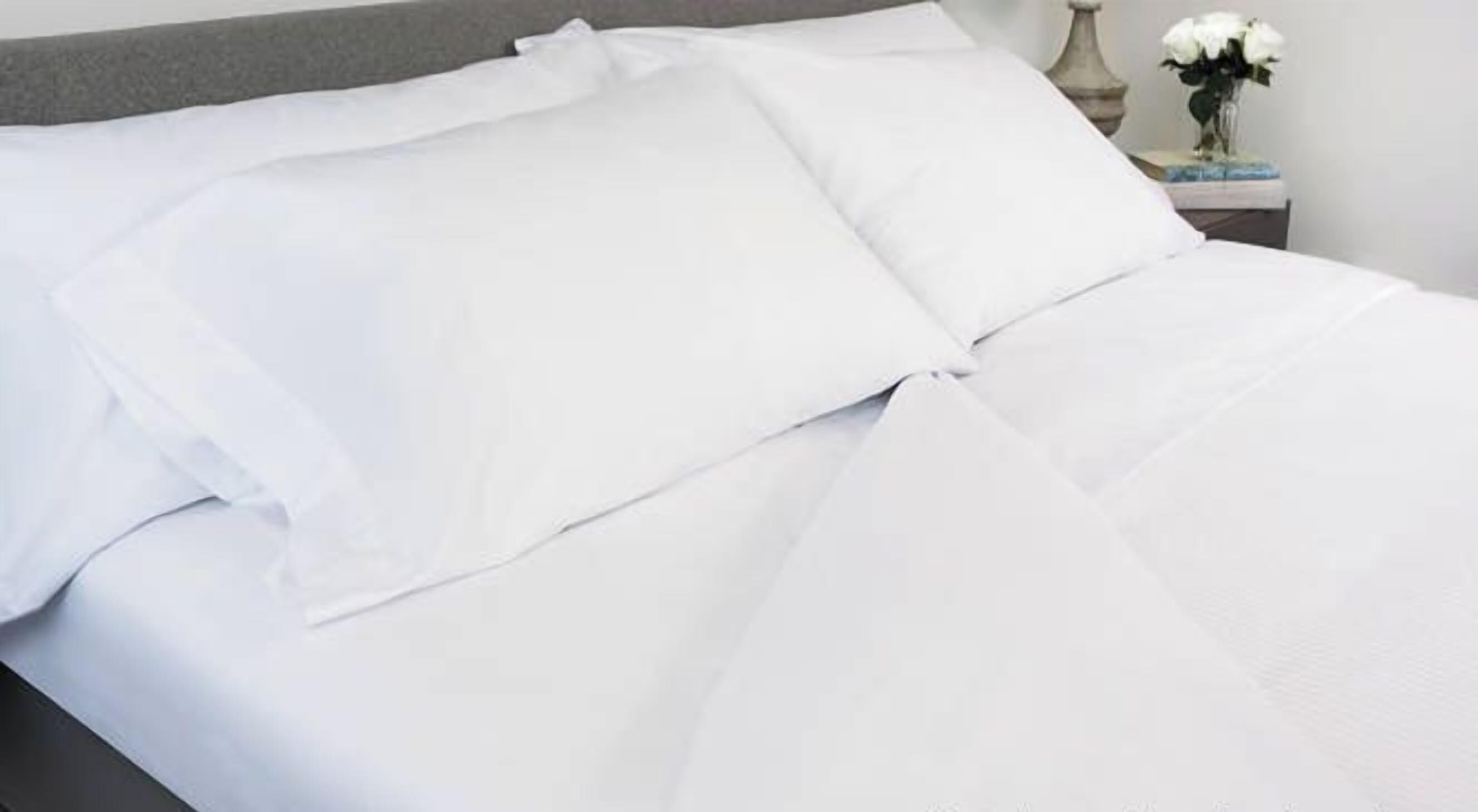 A neatly made bed with white bedding, a gray headboard, and a small bedside table with a vase of white flowers.