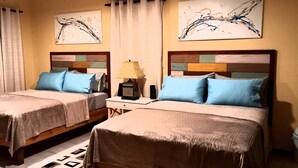 A hotel room with two beds, each with a wooden headboard and blue pillows. There is a small table with a lamp between the beds.