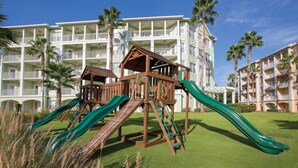 Ein Spielplatz mit Rutschen und Klettergerüsten vor einem Hotel mit mehreren Balkonen und Palmen.