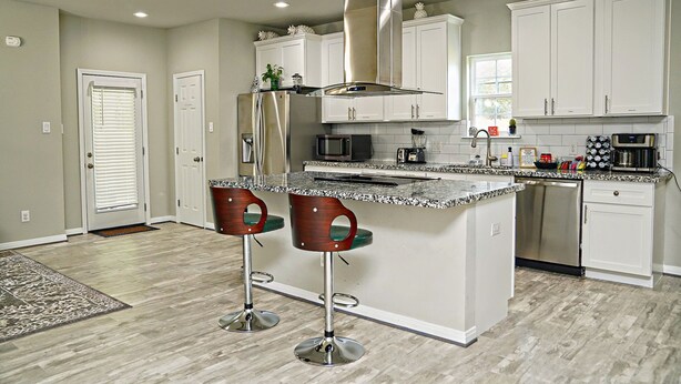 A modern kitchen with white cabinets, a stainless steel range hood, a granite countertop, and two bar stools. - Clean & Cozy new Home, 30 mins to George Bush INTL/ downtown 5 mins to woodlands (Conroe)