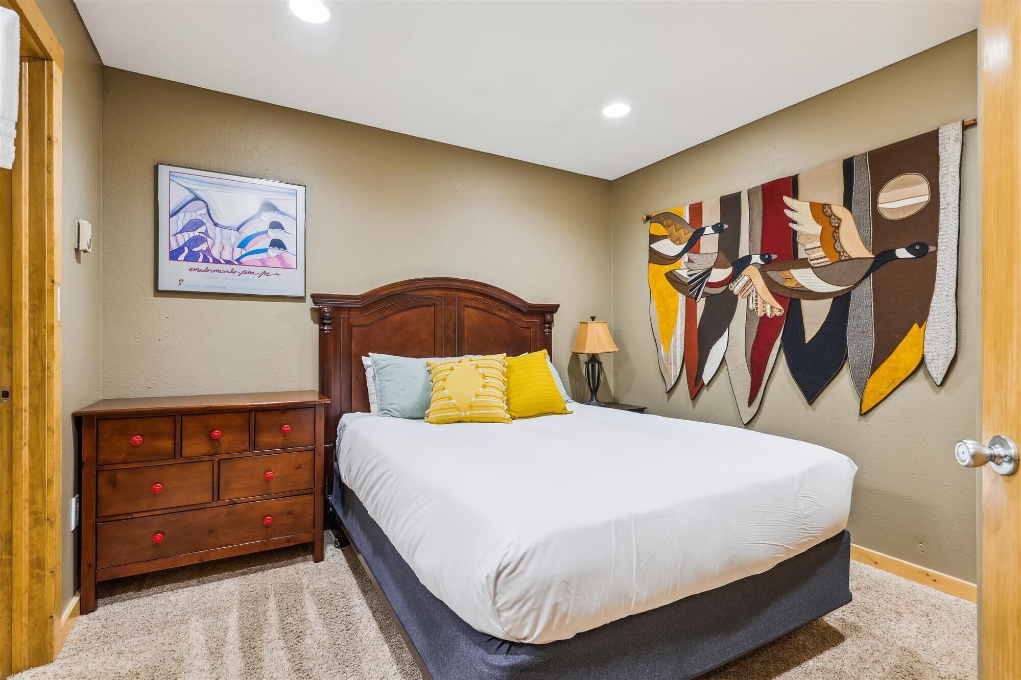 Un dormitorio con una cama de madera, una mesita de noche, un aparador con pomos rojos, una obra de arte en la pared y una puerta.