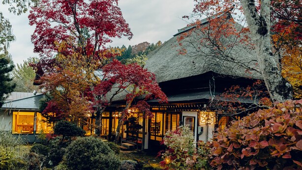 A traditional Japanese-style house with a thatched roof, surrounded by autumn foliage. - Takei Ryokan (Nagano)