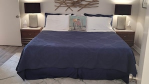 Un lit bien fait, recouvert d’une couvre-lit bleu foncé, avec deux lampes de chevet, une œuvre d’art abstraite accrochée au mur et un tapis à motifs posé sur un plancher en bois.