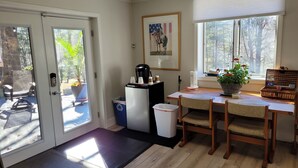 Une pièce comprenant une table à manger en bois, des chaises, un réfrigérateur, une poubelle, une fenêtre donnant sur des arbres et une porte menant à l’extérieur.