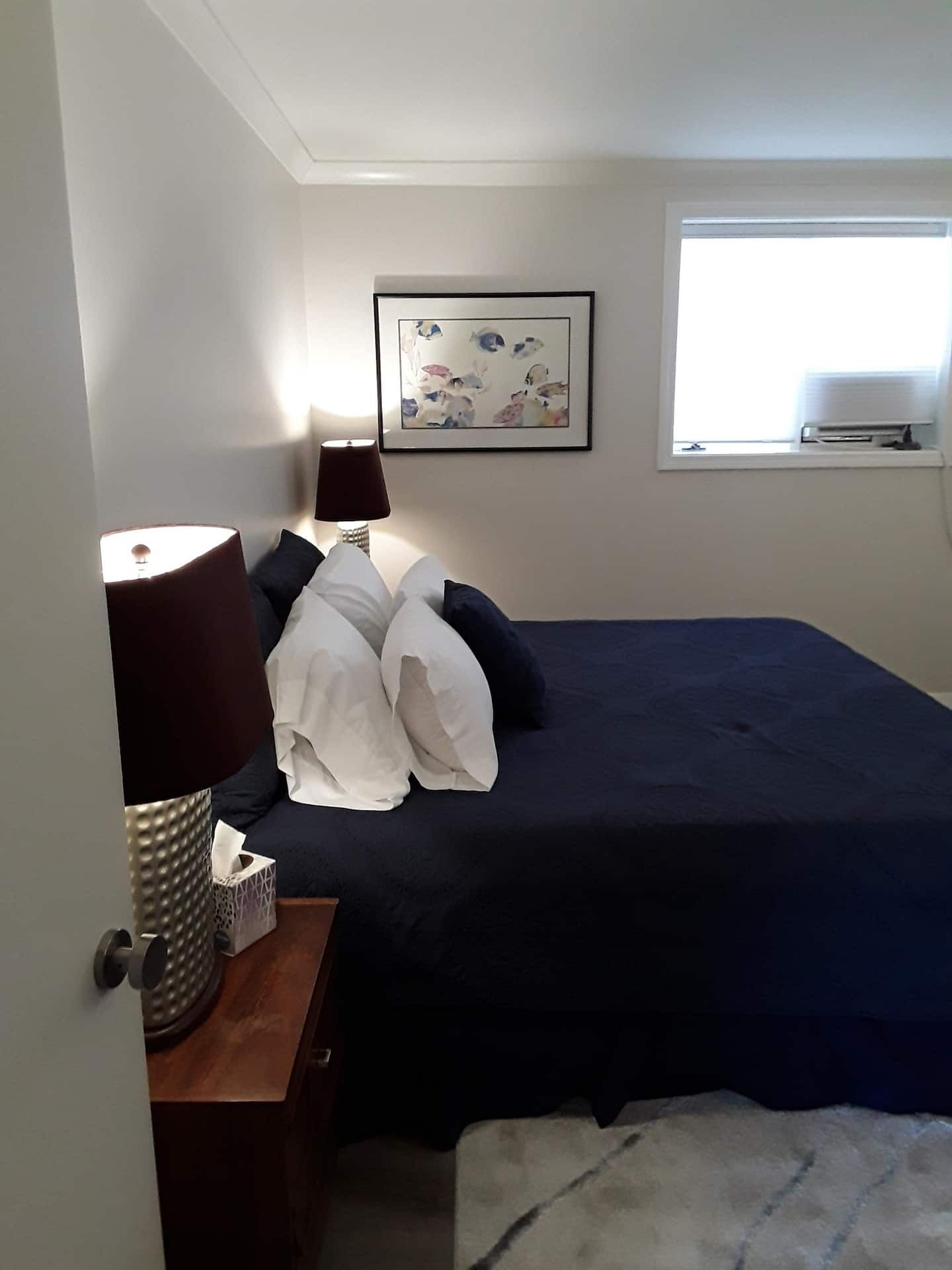 Un dormitorio con cama, mesita de noche con lámpara, un cuadro enmarcado en la pared y una ventana.